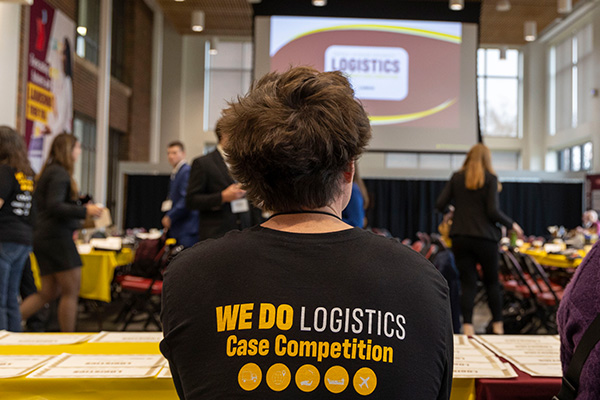 A student wearing a t-shirt which reads We Do Logistics Case Competition is prominently displayed in the foreground of a picture taken inside of Grawn's Atrium.