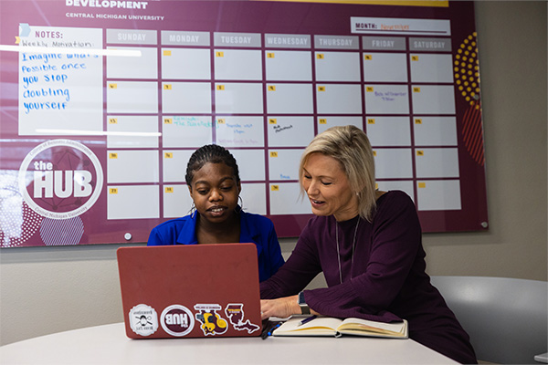 Lori Driessnack is helping a student in the Hub. They are both seated and looking into a laptop. Lori and the student are smiling.