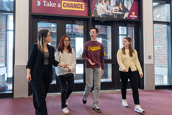 A small group of students are entering Grawn Hall's atrium through it's parking lot entrance doors. They are accompanied by a professor. They are dressed in business casual clothing.