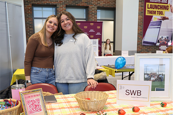 Two members of the Society of Women in Business are standing and posing behind their recruiting table at an event. There are several strawberry like decorations on the table.