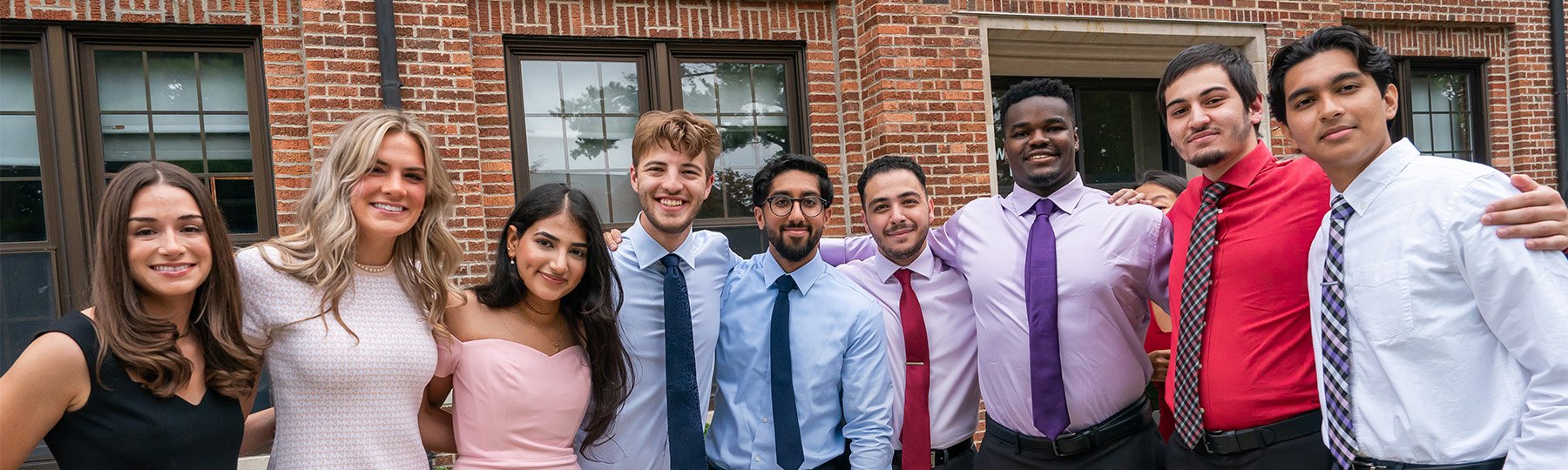 A diverse group of first-year medical students smiling and standing in a row with their arms around each other. A brick building is in the background.