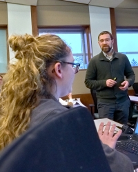 Conway_2019_200x250 A man in a dark sweater stands at the front of a classroom holding a presentation clicker and engaging with students. A woman with blonde hair types on a laptop. Large windows fill the bright classroom with natural light.