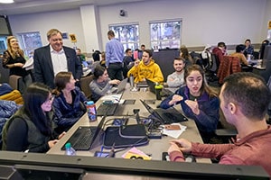 Students engage in a discussion while seated at a table with laptops.