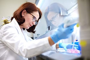 A woman wearing a white lab coat and blue gloves uses a pipette in a lab.