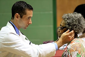 Man in a white lab coat inspects the ears of an older African American woman.