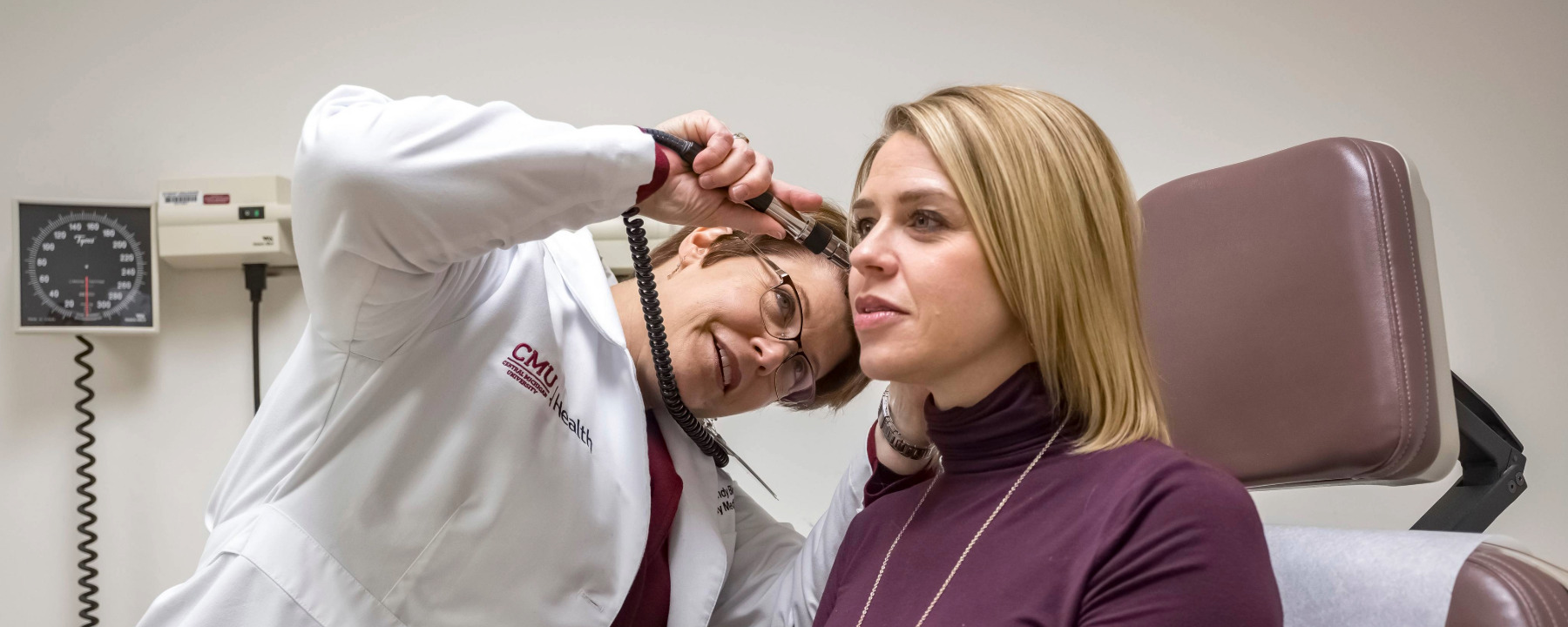 Careers_Biggs_1800x540 Wendy Biggs, M.D., in a medical exam room, using an otoscope to look in a female patient's ear.
