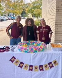 OSA_2025_Sag_200x250 Three people stand behind a table with a colorful plastic pool filled with rubber ducks. The table holds cups, snacks and promotional items. They wear matching maroon and gold attire. A brick building and trees are in the background.