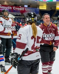 Hockey players wearing maroon and white jerseys with “Shocks and Saves” logos stand and talk on an ice rink. One player in the foreground wears jersey number 25. Bright arena lights and a credit union advertisement are visible in the background.