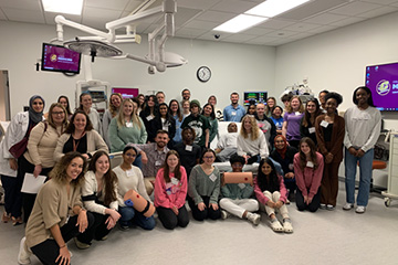 A large group of young students in a simulated hospital room, smiling at the camera.