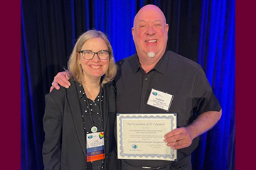 A woman and a man holding a certificate stand in front of a blue curtain smiling at the camera.