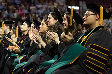 Graduates in medical school regalia are seated in a row and clapping at commencement.