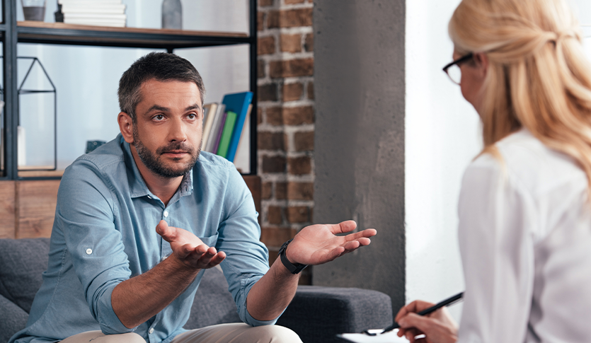 A man with a serious expression sits on a sofa, gesturing with his hands open, facing a woman with blonde hair in a white shirt who is holding a pen. They appear to be in a counseling session in a modern office with exposed brick walls.
