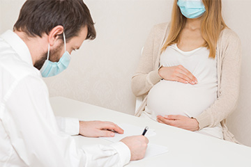Male doctor in a mask takes notes during an appointment with a pregnant patient.