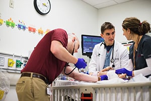 A young man in a white lab coat works with two others in an infant simulation lab.