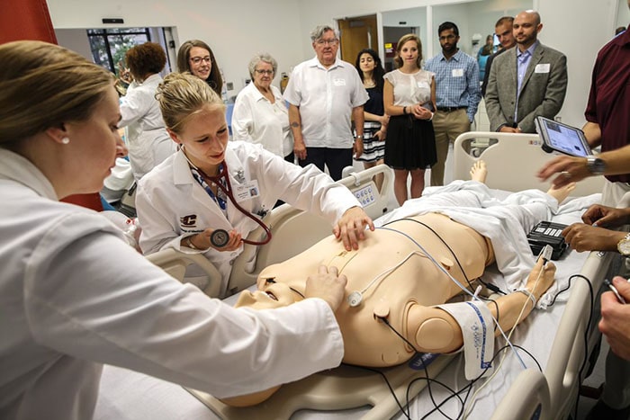 20210921_Saginaw_opening_simulation_700x467 People observe as two young women in white lab coats inspect a patient simulator.