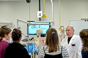 An older man in a white lab coat stands next to a skeleton and talks to students.