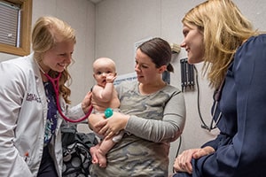 A young woman in a shite lab coat smiles as she uses a stethoscope to examine a baby being held by another woman.