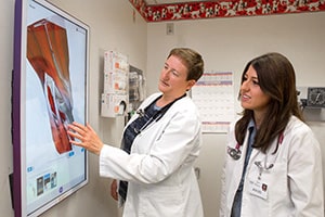 A woman in a white lab coat points out anatomy on a screen to a younger woman in a white lab coat.