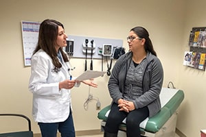 A young woman in a white lab coat holds a paper in one of her hands while she speaks to a woman sitting on an examination table.