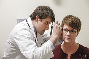 A young man in a white lab coat inspects the ear of a woman.