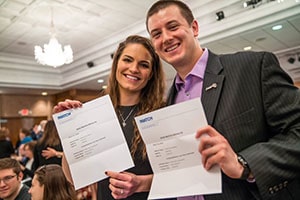 An young man and a young woman stand together and smile while holding letters detailing their Match Day assignments.