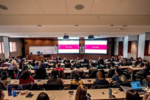 A large group of students sits in a lecture room and looks towards a screen.