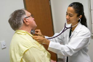 A medical student in a white coat listens to an older patient's heartbeat with a stethoscope. The patient, wearing a yellow shirt, sits on an examination table in a medical office.
