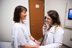 A medical student is smiling and using an otoscope to examine a female patient's ear in a medical office. The patient is wearing a hospital gown and sitting on an examination table.