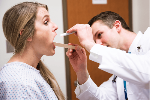 A medical student examines a patient's throat using a tongue depressor and a light. The patient is in a hospital gown and seated, while the medical student focuses on the procedure.