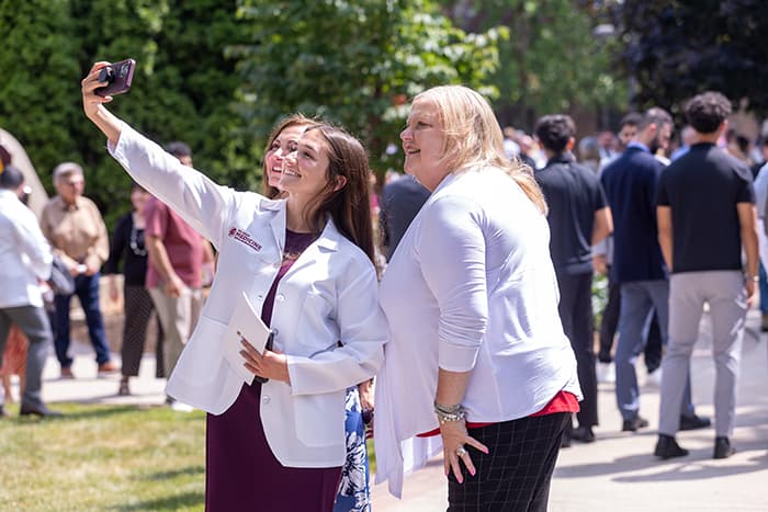 2025_CMEDWhiteCoat_700x467 Three women are taking a selfie outdoors during a sunny day. People and greenery are visible in the background.