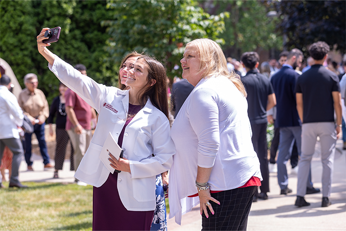 Three women are taking a selfie outdoors during a sunny day. People and greenery are visible in the background.