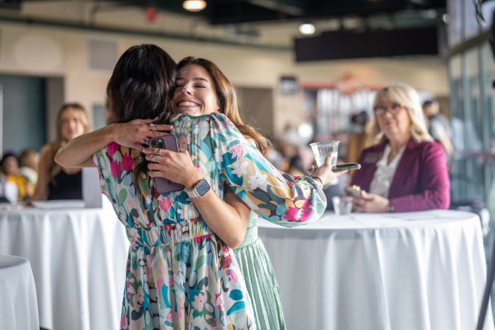 A medium shot shows two young women joyfully hugging at an indoor event. One, facing the camera, smiles and wears a colorful floral dress. The other, seen from behind, wears a matching dress. Other attendees are visible in the softly blurred background.