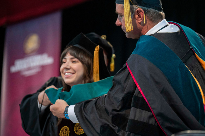 A faculty member in academic regalia places a green doctoral hood over a smiling female graduate’s head during a hooding ceremony. Both wear black caps and gowns with gold tassels. A blurred maroon banner is in the background.