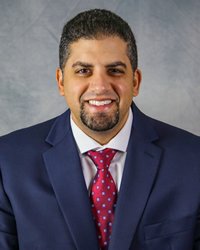 A man with dark hair and goatee wears blue suit with white collared shirt and red tie.