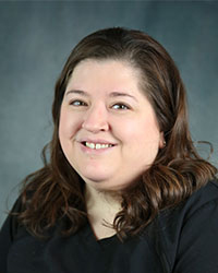 a woman with brown hair wearing a gray shirt smiling and standing in front of a gray background