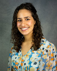 A woman with curly hair wearing a floral shirt smiles for a professional headshot against a grey background.