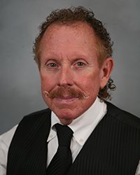 A professional headshot of Ronald Bradley in black and white attire against a gray background.