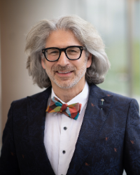 A man with gray hair and a beard, wearing dark-framed glasses and a colorful bow tie, is dressed in a dark suit with a white shirt. The background is softly blurred, suggesting an indoor setting.