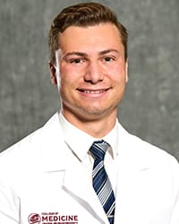 A man in a white button up, tie and medical white coat smiles for a head shot.