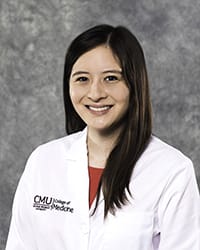 A woman with long, dark hair and a red blouse wears a white coat and smiles for a headshot.