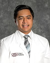 A man with brown hair wears a white collared shirt, tie and medical white coat in a headshot image.
