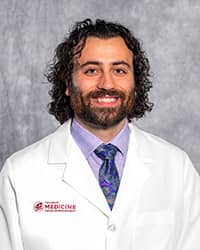 A man with medium-length curly brown hair wearing a lavender shirt, multi-color tie and a medical white coat as he smiles at the camera.