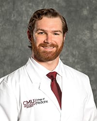 A man with a beard wears a white button up, red tie and white medical coat in a headshot image.