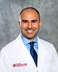 A man wearing a blue shirt, tie and a medical white coat as he smiles at the camera.