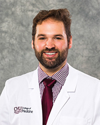 A man with short brown hair, full beard and mustache wearing a maroon checked shirt, maroon tie, and a medical white coat as he smiles at the camera.