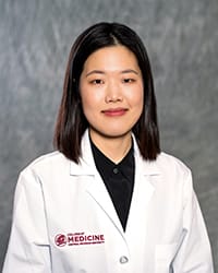 A woman with black hair and a black blouse wears a white coat and smiles for a headshot.