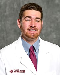 A man with a beard wears a grey button up, red tie and white medical coat in a headshot image.