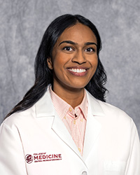 A woman with long black hair wearing a pink shirt and a medical white coat as she smiles at the camera.