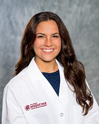 A woman with long, dark hair and a blue blouse wears a white coat and smiles for a headshot.