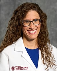 A woman with curly, brown hair and a blue blouse wears a white coat and smiles for a headshot.